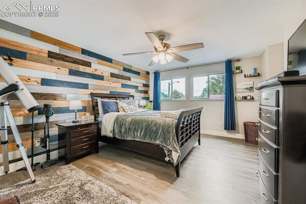 Image 12 of 27: Bedroom with light wood finished floors, wood walls, and a ceiling fan