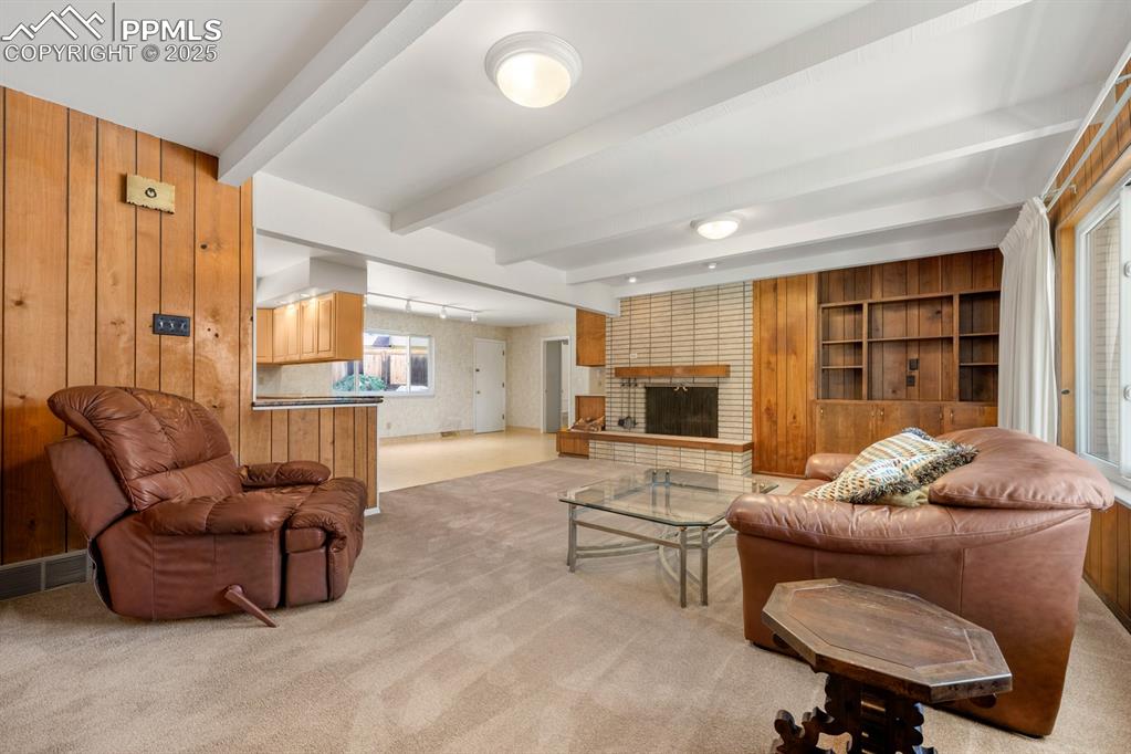 Image 6 of 50: Living room with wood walls, light carpet, beam ceiling, and a fireplace