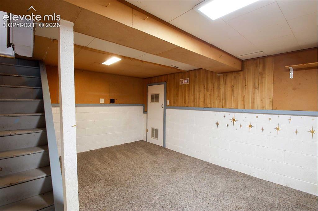 Image 17 of 19: Carpeted flooring, painted cinder block walls, wood paneling, stair access,