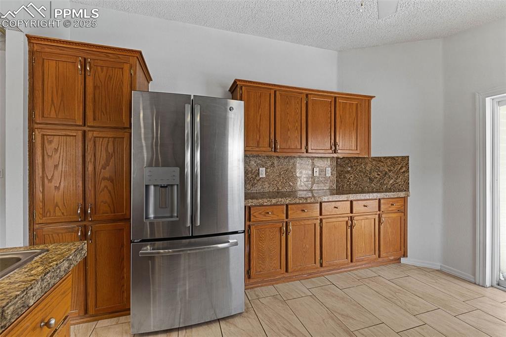 Image 7 of 24: Kitchen with stainless steel fridge, brown cabinetry, dark countertops, and
